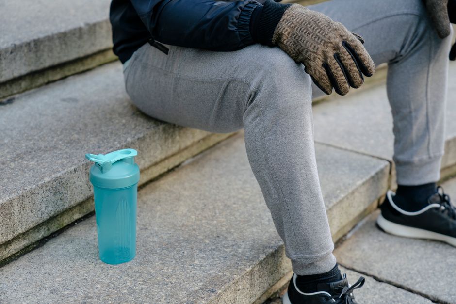 Crop anonymous male in gray sweatpants and sneakers and gloves sitting on steps of stairway with plastic fitness bottle